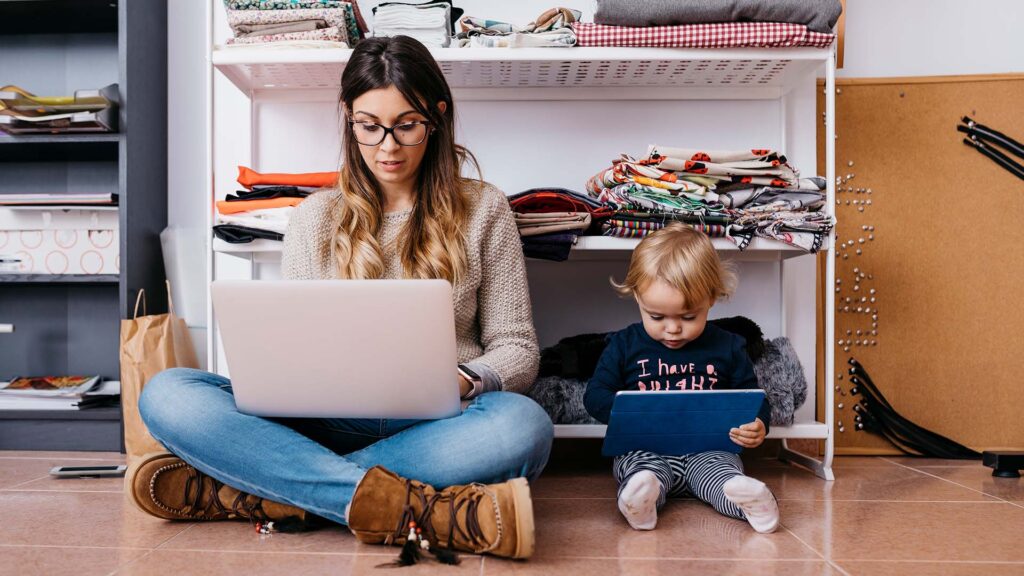 Adult working remotely on laptop sitting next to child holding a tablet