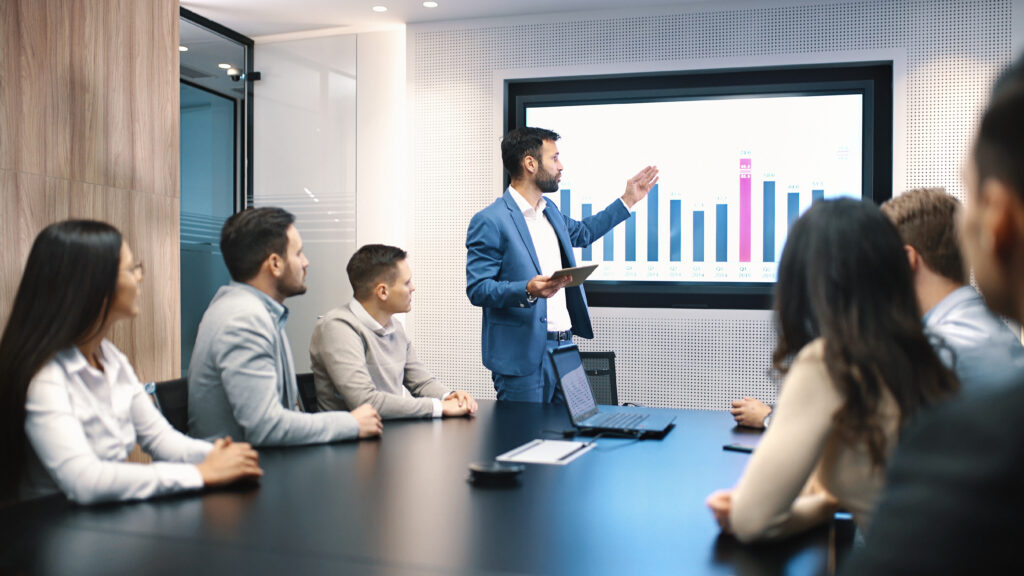 Meeting in board room with one person presenting a bar graph of financials to colleagues around a large desk