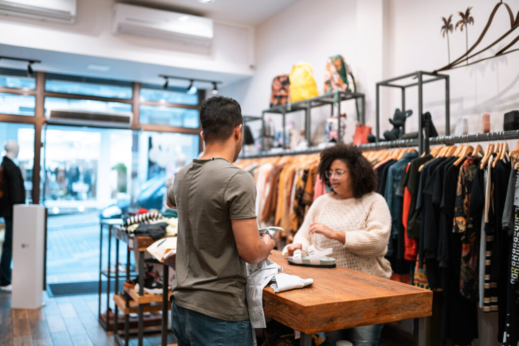 An attendant in a clothing retail store helping a customer to find clothes and shoes. The attendant has their back turned and the customer is holding a pair of white sandals on the wooden table between them.