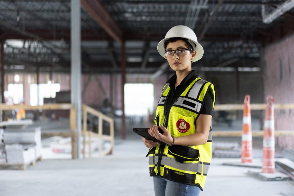 Construction worker wearing a yellow hardhat and safety vest, holding a tablet in a warehouse