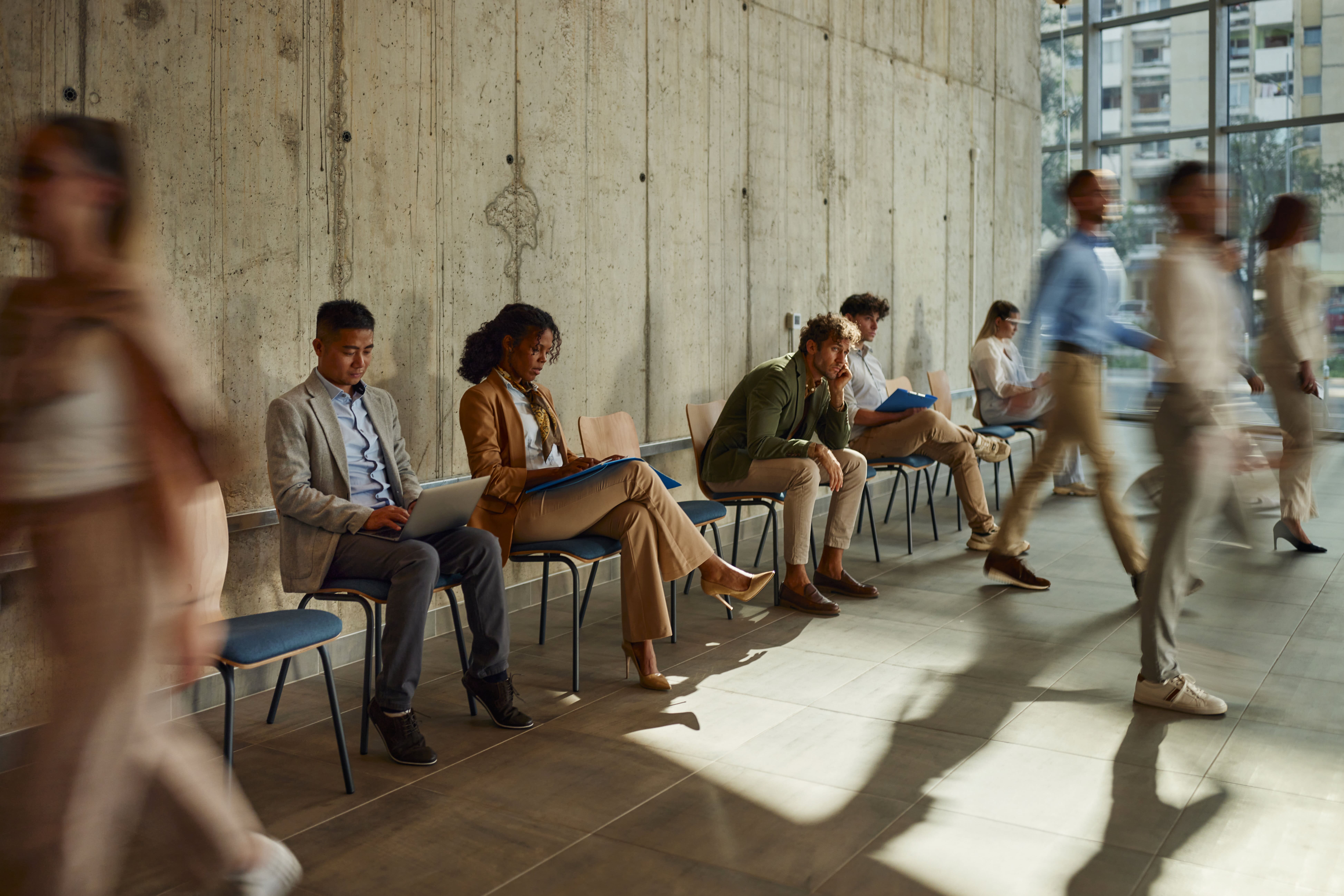 People sitting in a building lobby waiting for job interviews. Some are seated and clear while others are walking and thus are blurred.