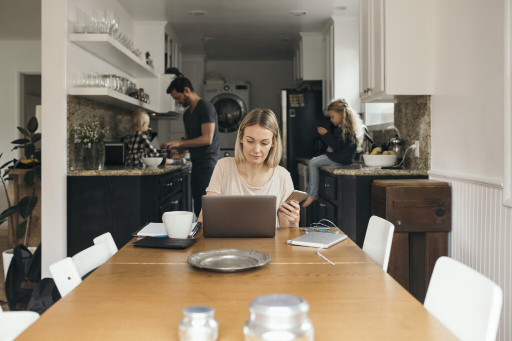 Person holding phone in front of laptop working from the kitchen table with their spouse and child in the kitchen behind them.